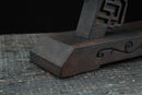 Supporto per katana in legno scuro, vista angolata laterale, intagli elaborati e finitura liscia, su una superficie rustica. Supporto Katana Douu