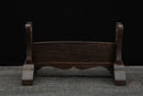 Supporto per katana in legno scuro con gambe curve e schienale piatto, su sfondo nero e superficie rustica. Supporto Katana Keshiki.