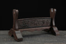 Supporto per katana in legno marrone scuro con intagli elaborati sul pannello frontale, rialzato su una superficie testurizzata. Supporto Katana Keshiki.