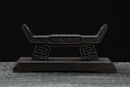Supporto per katana in legno scuro con intagli elaborati e dettagli decorativi, vista frontale su una superficie testurizzata. Supporto Katana Douu.