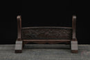 Supporto per katana in legno scuro con elaborati intagli floreali, visto di fronte su una superficie testurizzata. Supporto Katana Keshiki.