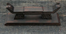 Supporto per katana in legno scuro decorato con intagli elaborati, visto frontalmente su una superficie testurizzata, Supporto Katana Douu.