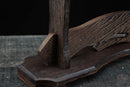 Primo piano di un supporto per katana in legno scuro con venature elaborate e dettagli curvi su una superficie rustica. Supporto Katana Otera.
